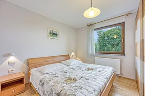Un dormitorio con cabecera de madera, un ventanal con vistas a árboles, una cama con colcha floral y lámparas de noche.