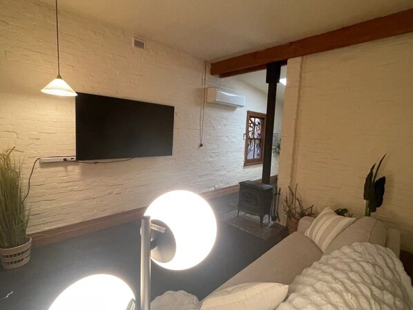 A living room with a flat-screen TV mounted on a white brick wall, a sofa, a coffee table, and a potted plant. - Cozy Bluestone House in Norwood – Perfect for Families and Their Pets (Norwood)