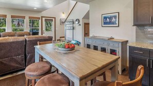 Un coin repas avec une table en bois, quatre chaises en cuir marron et un saladier rempli de fruits. Des fenêtres offrent une vue sur la verdure et un bâtiment situé à l’extérieur.