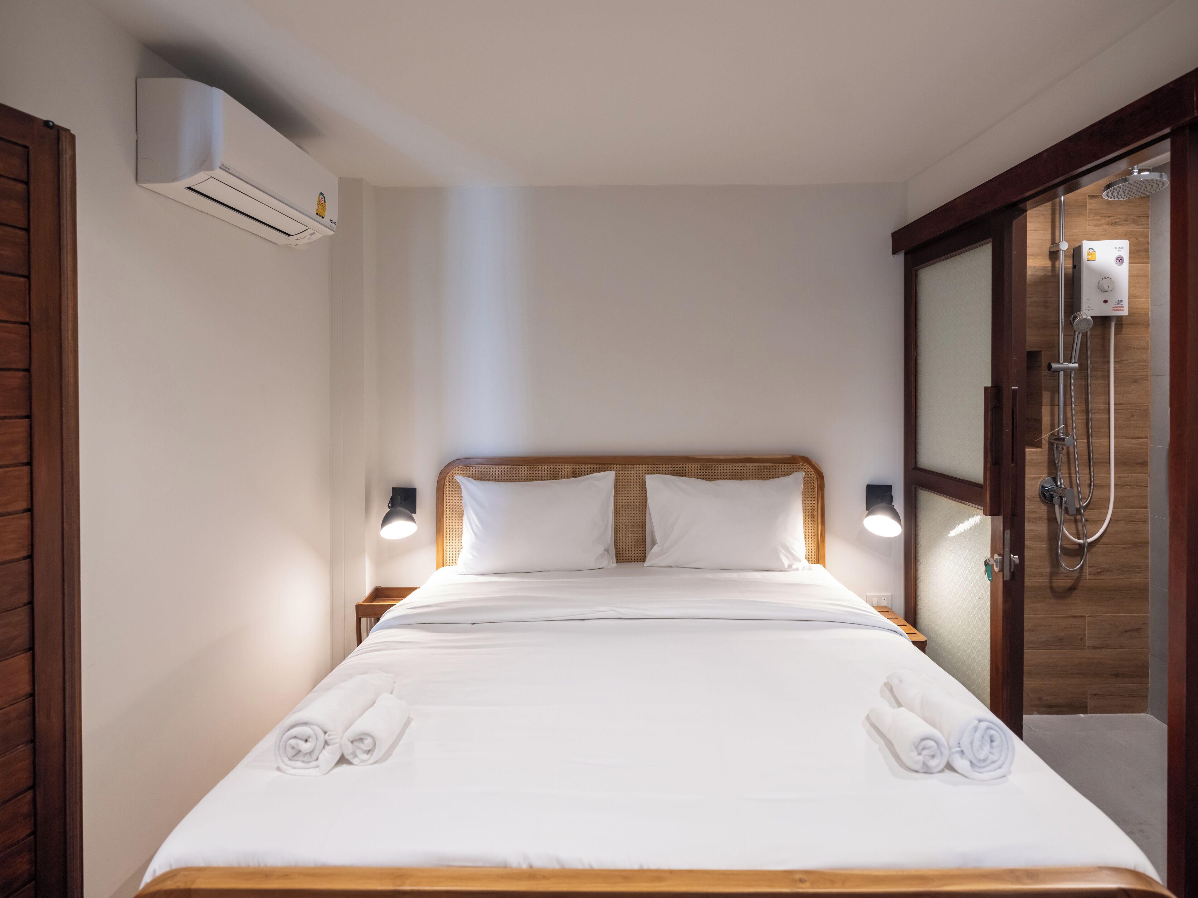 A hotel room with a wooden bed, white linens, two rolled towels, a wall-mounted air conditioner, and a shower area with a control panel.