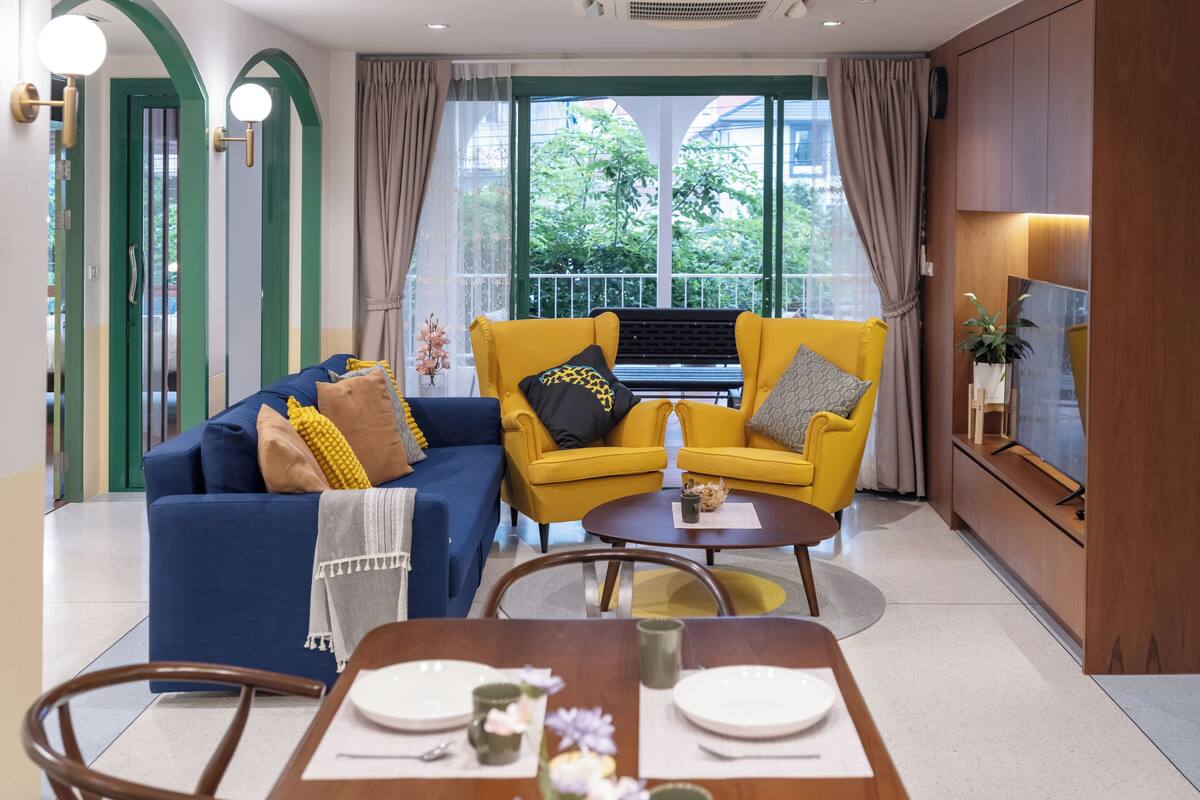 A modern living room with a blue sofa, yellow armchairs, a dining table set for four, and a large window with a view of greenery.