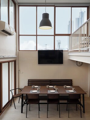 A dining area with a large window, a hanging light fixture, a flat-screen TV, and a clock on the wall. - Bedtime Storeys (Bangkok)