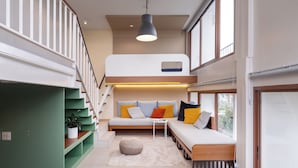 Un interior moderno con una escalera que lleva a una habitación estilo loft, un sofá con cojines y una lámpara colgante.
