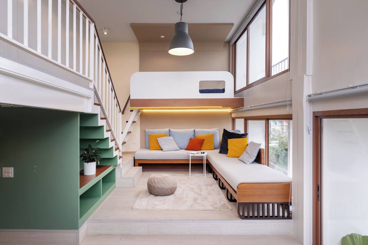 A modern interior with a staircase leading to a loft bedroom, a sofa with cushions, and a hanging light fixture.