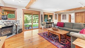 A spacious living room with a stone fireplace, wooden beams, a sofa, and a large window. - Mountain chalet w/ hot tub & games (Whistler)