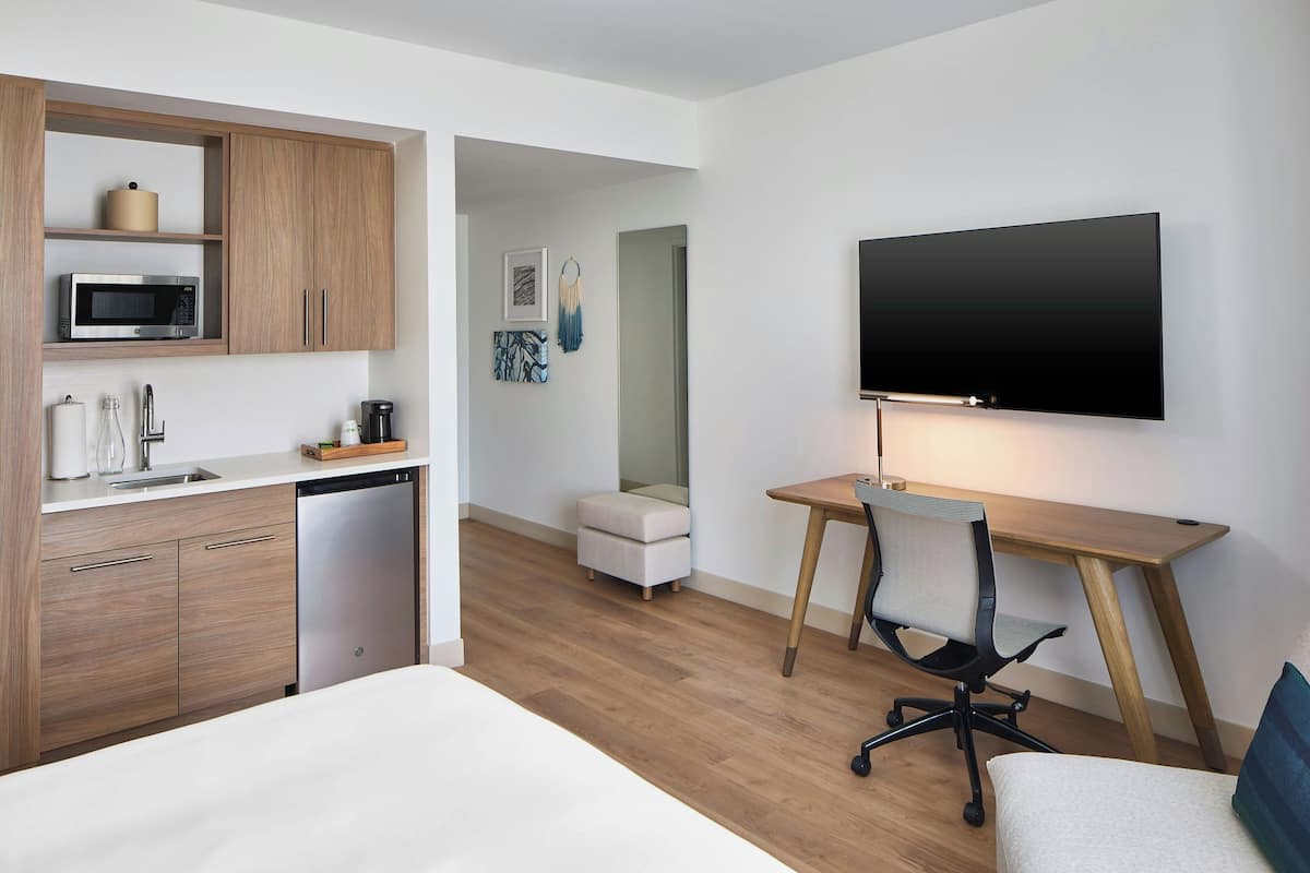 A modern hotel room with a kitchenette, a flat-screen TV, a wooden desk, a comfortable chair, a bed, and a small seating area.