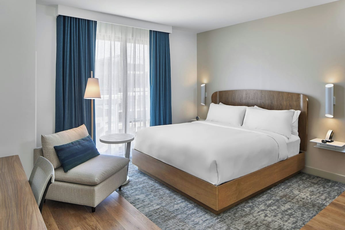 A modern hotel room with a large bed, a small armchair, a side table, and a view of the outdoors through sheer curtains.