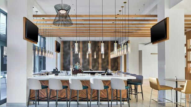 A modern bar area with a long counter, multiple bar stools, hanging pendant lights, and two flat-screen TVs mounted on the wall.
