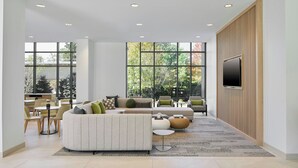 A modern living room with a sectional sofa, multiple seating options, a flat-screen TV mounted on a wooden panel, and large windows offering a view of the outdoors.