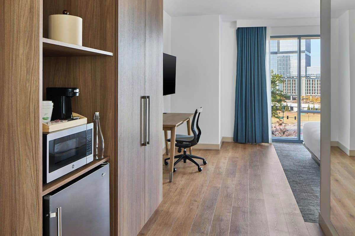 A modern kitchen with a microwave, coffee maker, and refrigerator. There is a dining table with a chair and a large window with curtains.