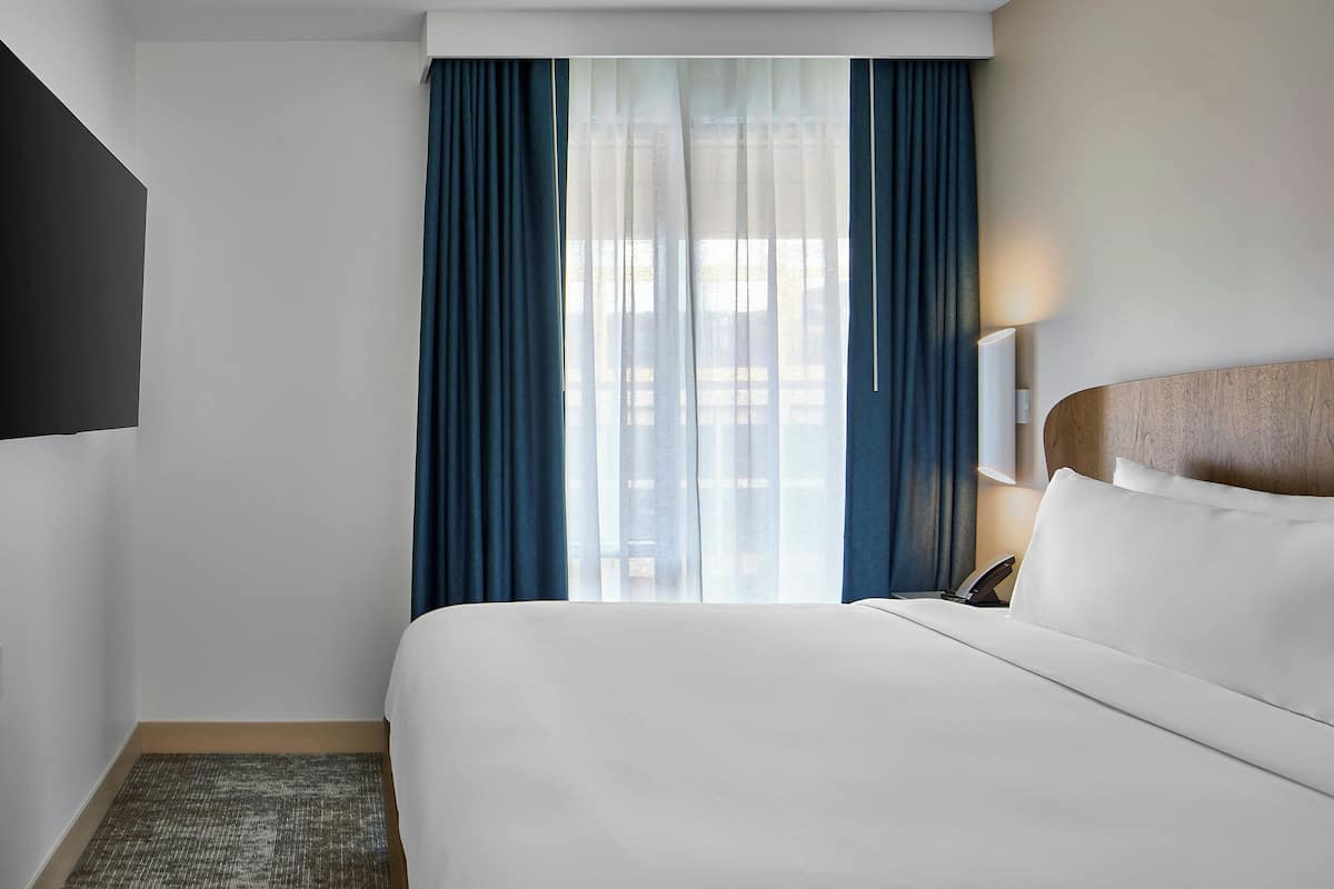 A hotel room with a bed, a TV mounted on the wall, a bedside table with a lamp, and a window with curtains.