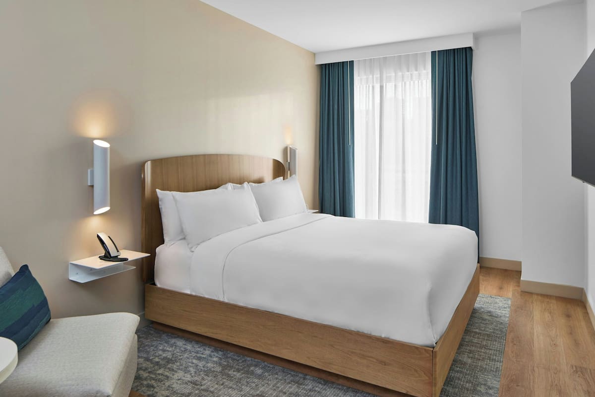 A modern hotel room with a wooden bed, a bedside table with a lamp, a small shelf, and a large window with curtains.