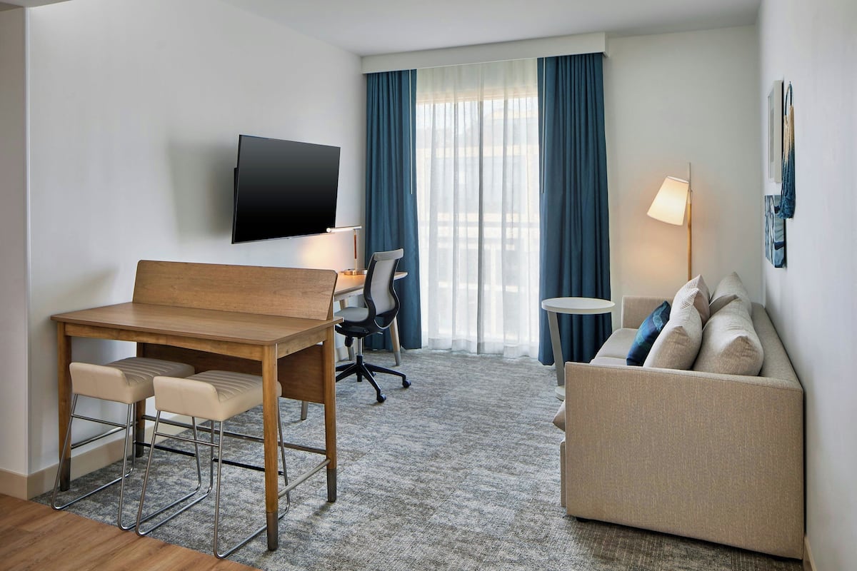 A hotel room with a wooden desk, two chairs, a sofa, a TV, a lamp, and a window with curtains.