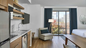 A modern kitchen with a built-in dishwasher, a dining area with a wooden table and chairs, a sofa, and a large window with a view of a building and trees.