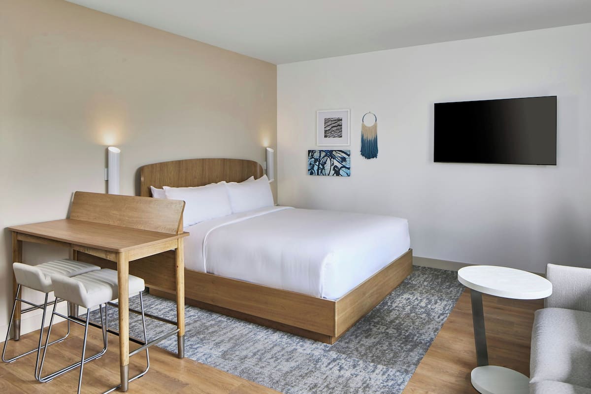 A modern hotel room with a wooden bed, a small desk, two chairs, and a round table.
