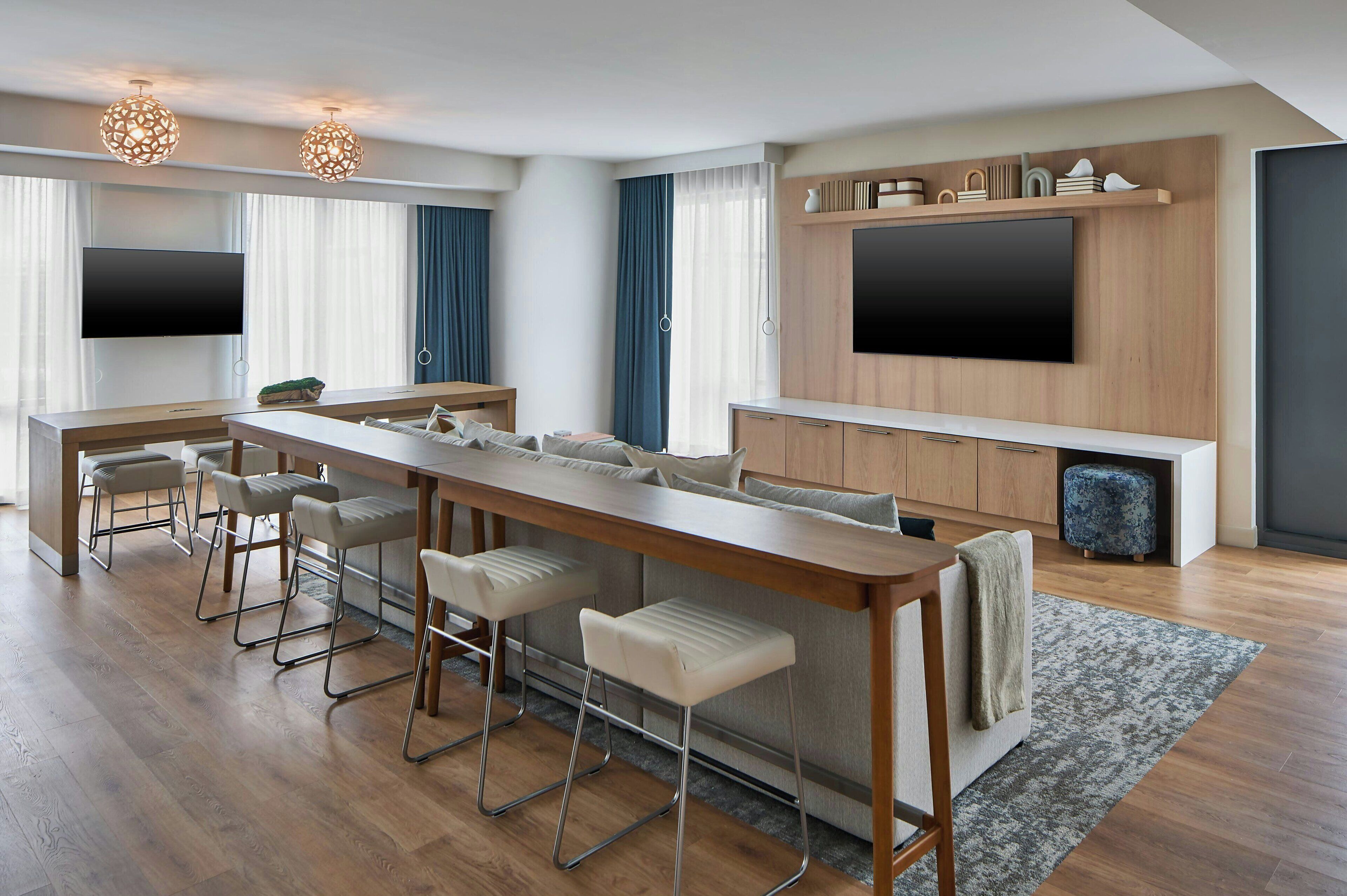 A modern living room with a long wooden table, bar stools, a flat-screen TV mounted on the wall, and a large window with curtains.