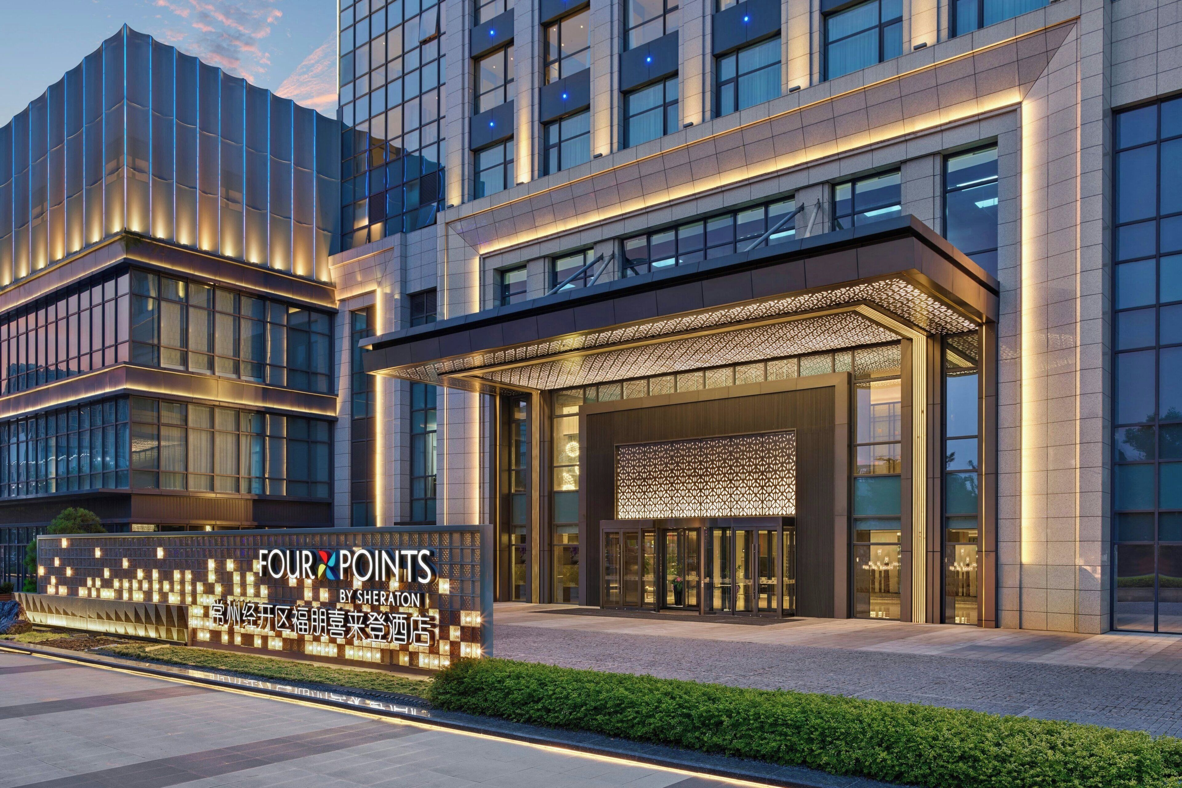 A modern hotel entrance with a large illuminated sign, glass doors, and a covered entrance area.