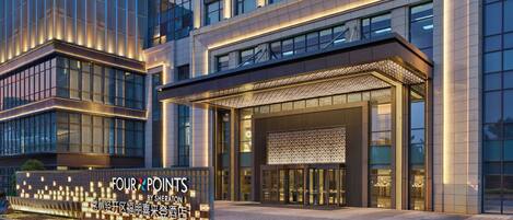 A modern hotel entrance with a large illuminated sign, glass doors, and a covered entrance area.