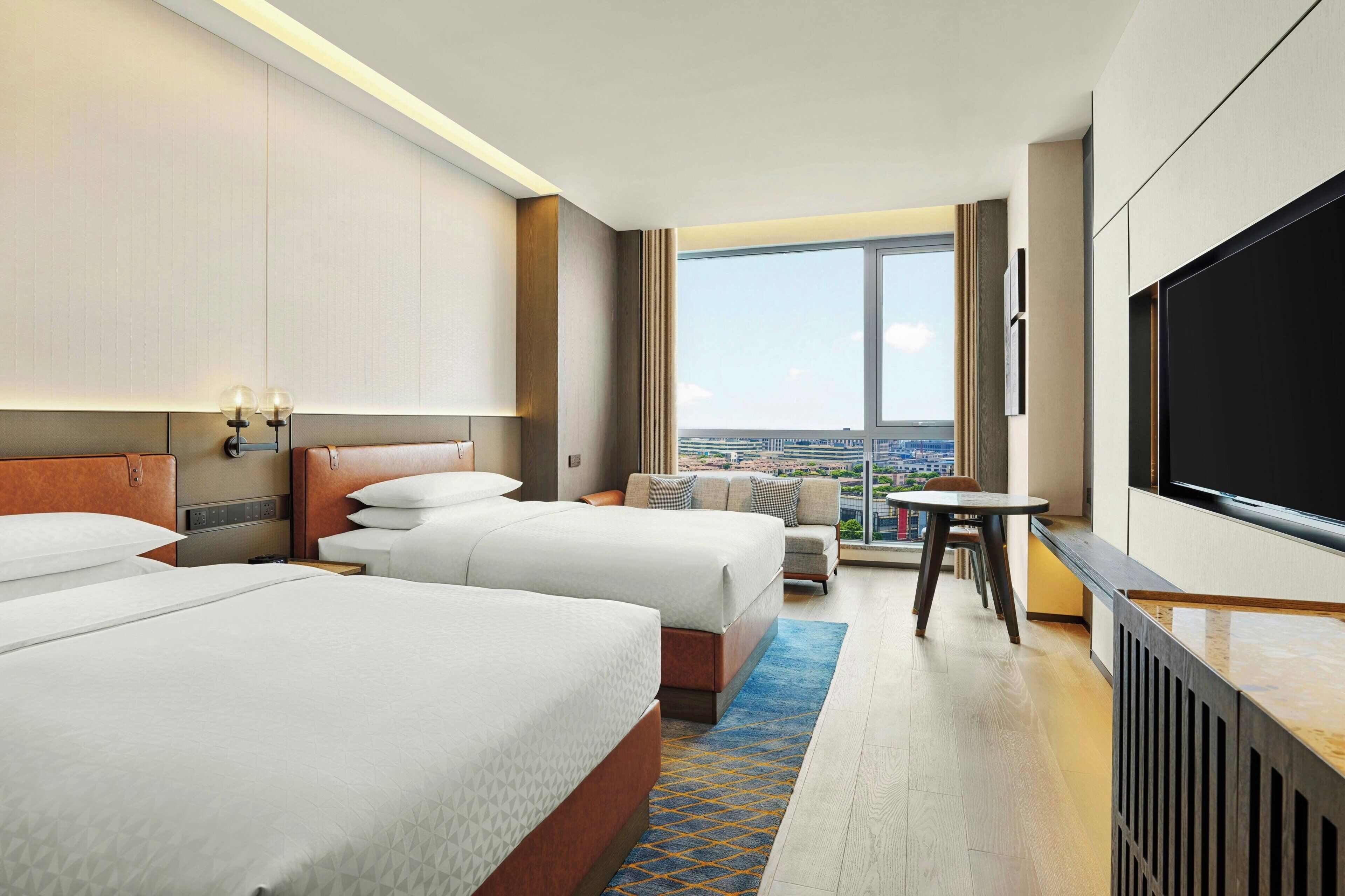A hotel room with two beds, a sofa, a TV, and a view of the city.