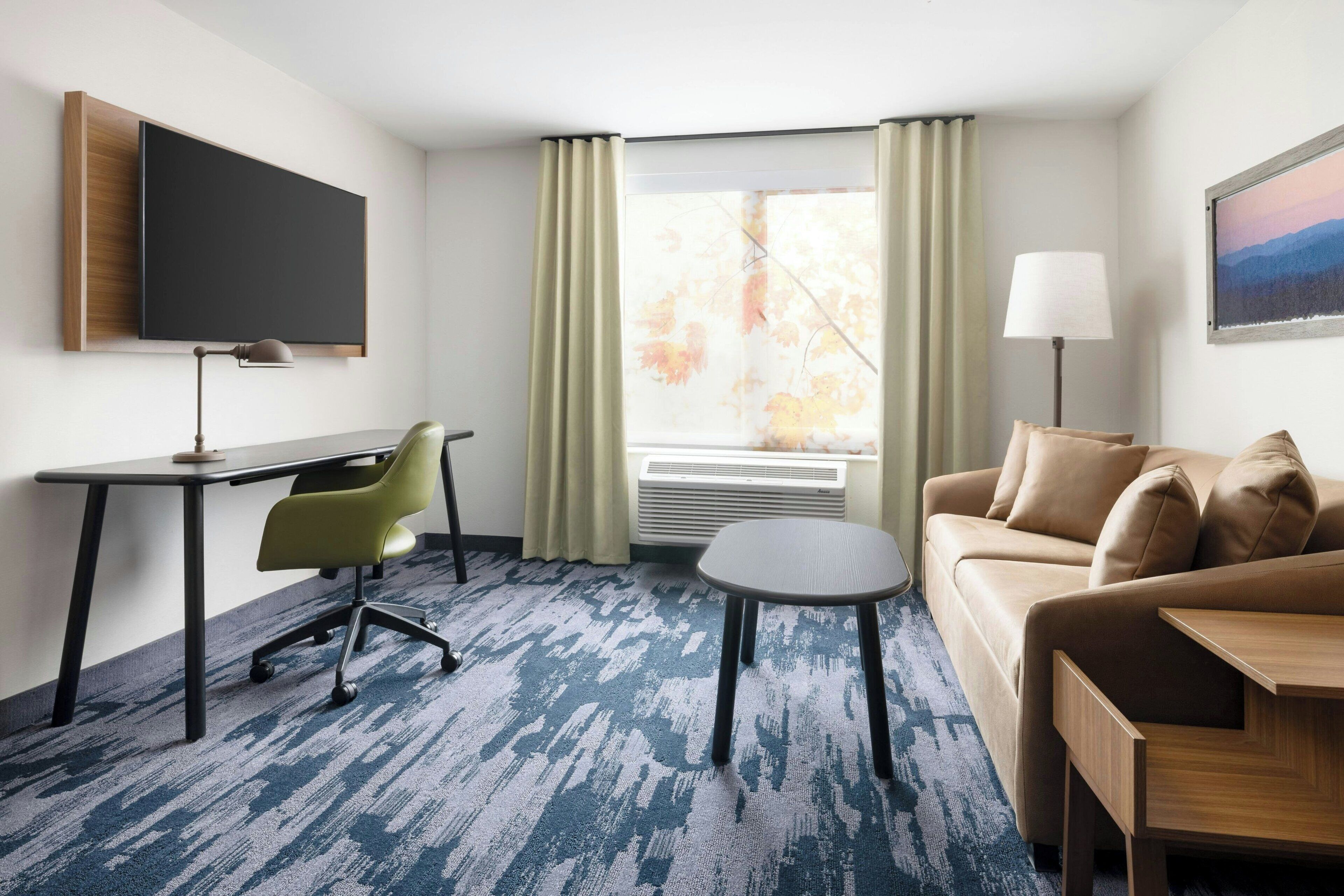 Une chambre d’hôtel équipée d’un bureau, d’un fauteuil de bureau vert, d’une petite table ronde, d’un canapé, d’un téléviseur fixé au mur, d’une fenêtre avec des rideaux et d’un appareil de climatisation.