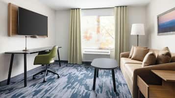 A hotel room with a desk, green office chair, a small round table, a sofa, a TV mounted on the wall, a window with curtains, and an air conditioning unit.