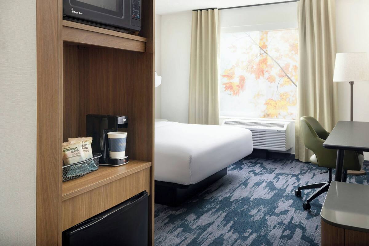 A hotel room with a bed, a desk, a chair, a microwave, a coffee maker, and a window with curtains.