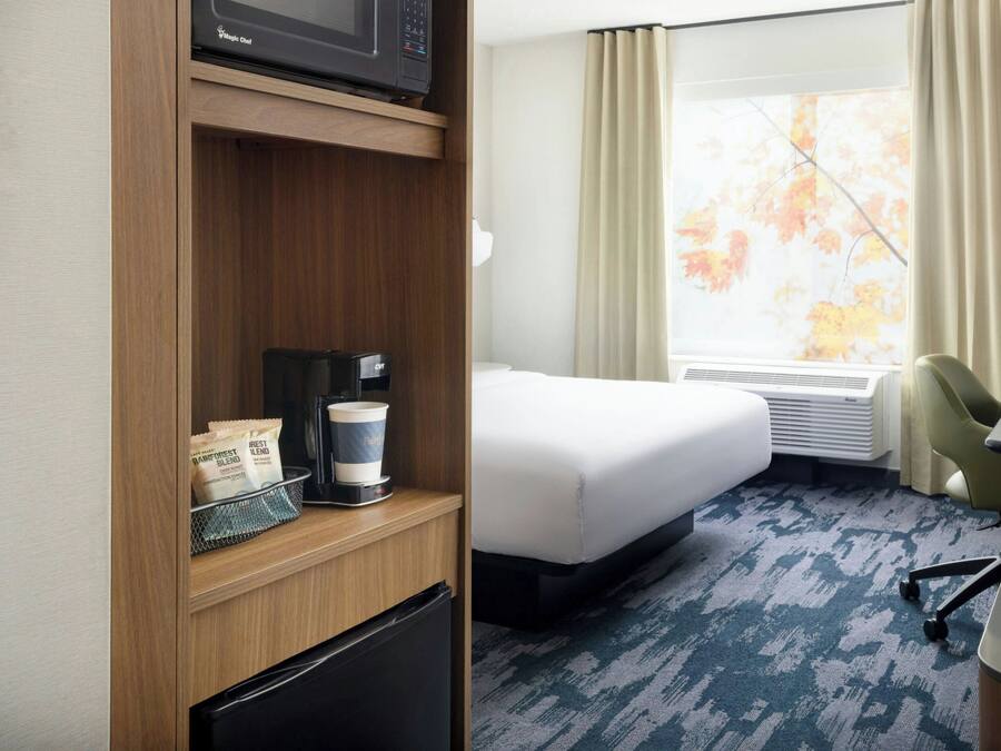 A hotel room with a bed, a desk, a chair, a microwave, a coffee maker, and a window with curtains.