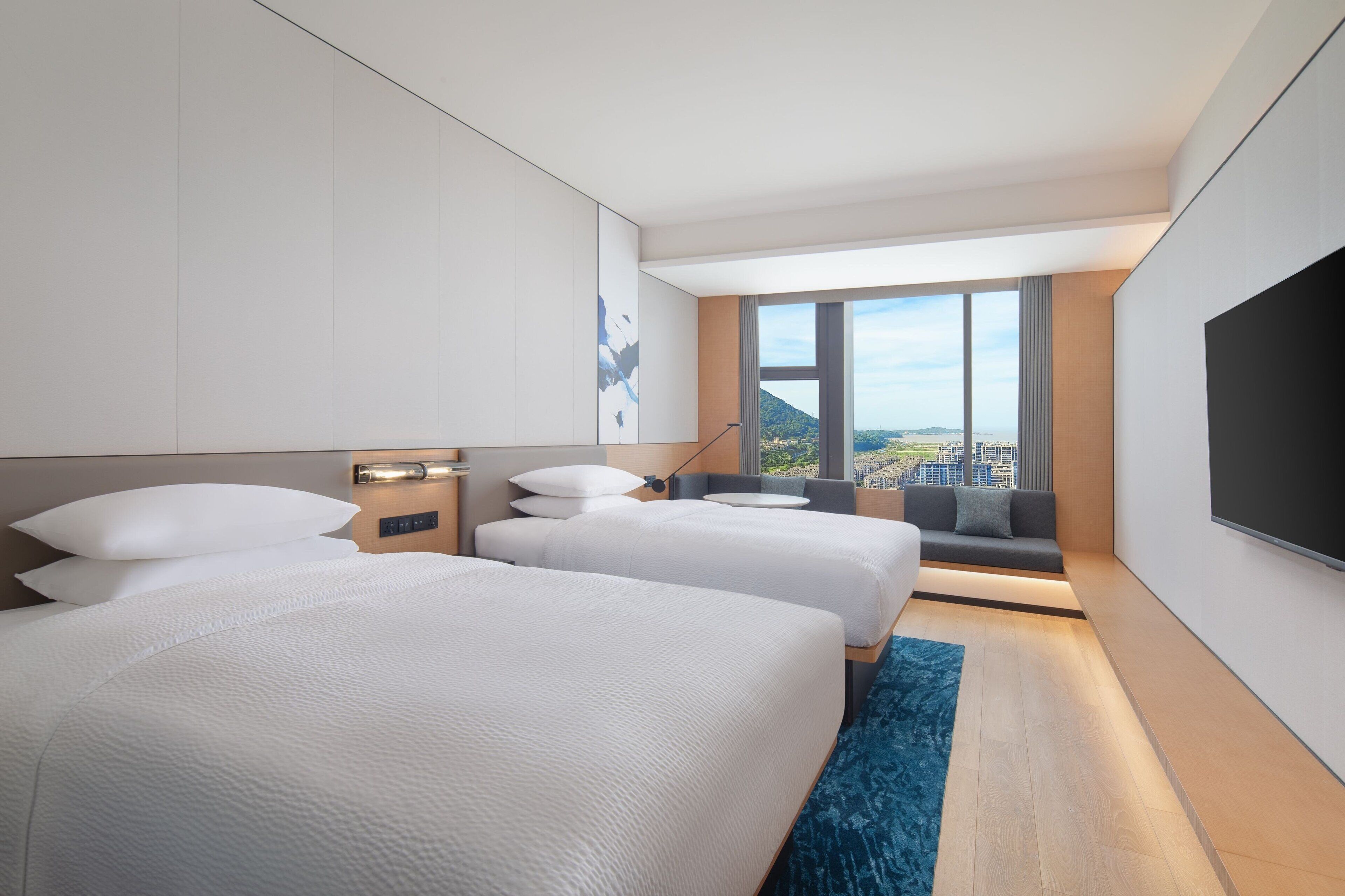 A modern hotel room with two beds, a large window offering a city view, a flat-screen TV, and a small seating area.