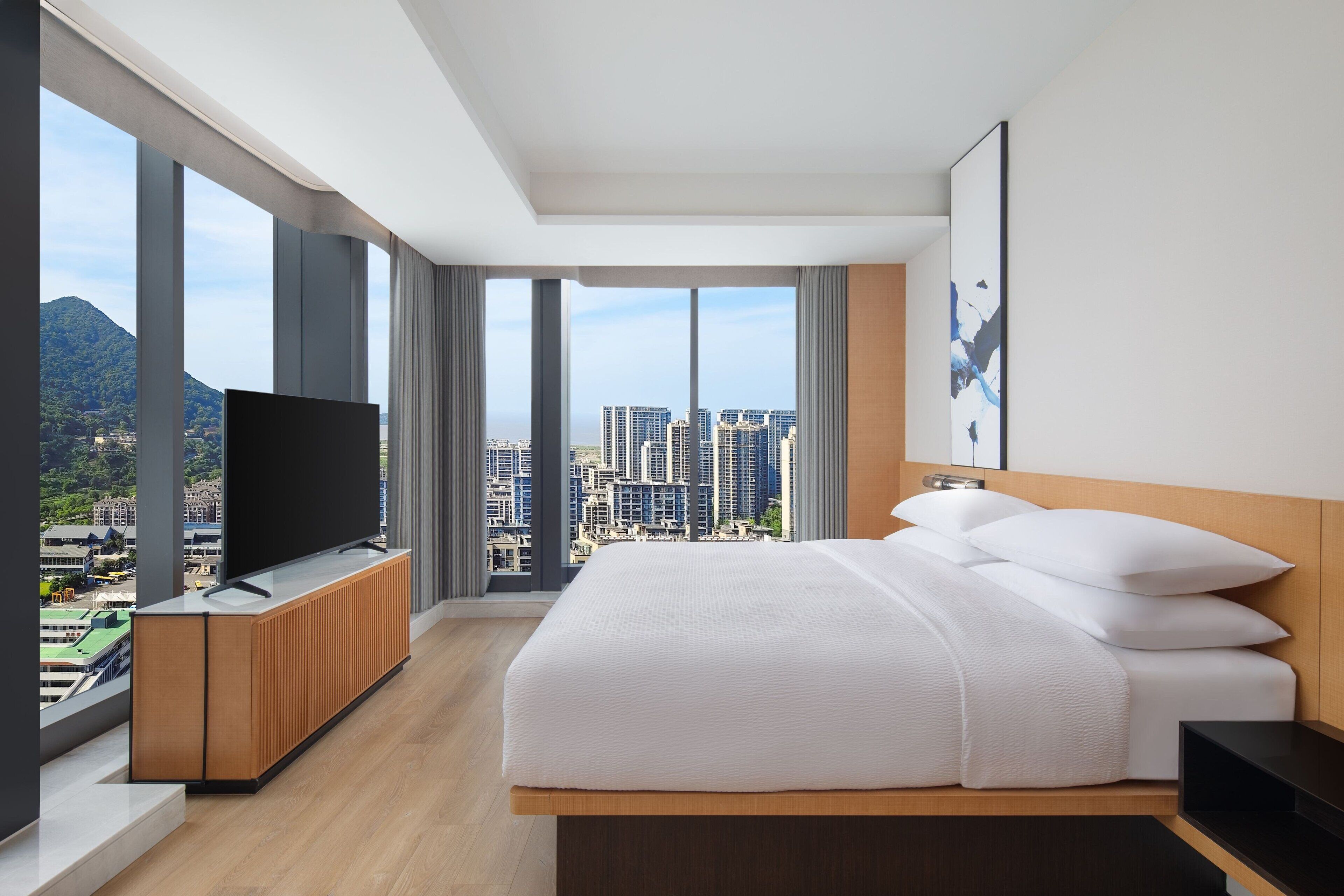 Habitación de hotel moderna con una cama grande, un televisor de pantalla plana y vistas a la ciudad a través de amplios ventanales.