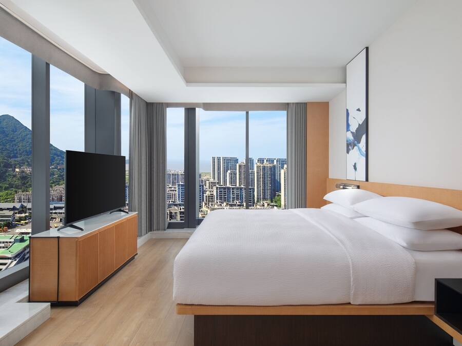 Ein modernes Hotelzimmer mit einem großen Bett, einem Flachbildfernseher und Blick auf die Stadt durch große Fenster.