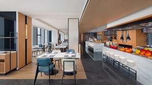 A modern dining area with long tables and chairs, a kitchen counter with food items, and a bar area with stools.