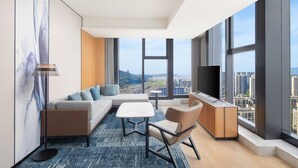 A modern hotel room with a large window, a sofa, a coffee table, a chair, and a TV.