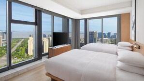 A modern hotel room with large windows, a flat-screen TV, a wooden nightstand, and a bed with white bedding.