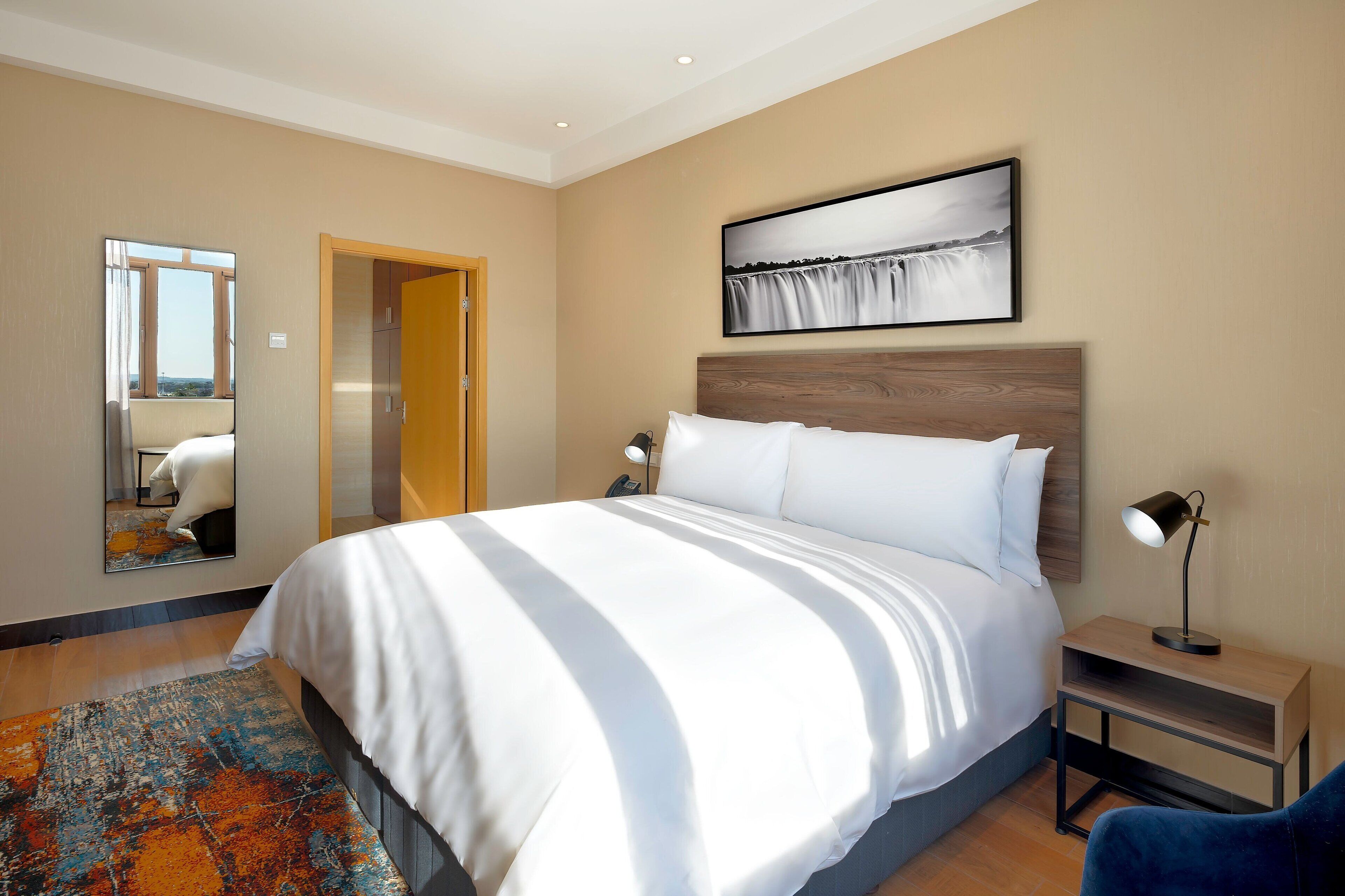 Habitación de hotel moderna con una cama grande, una mesita de noche con lámpara, un espejo y un cuadro en la pared.