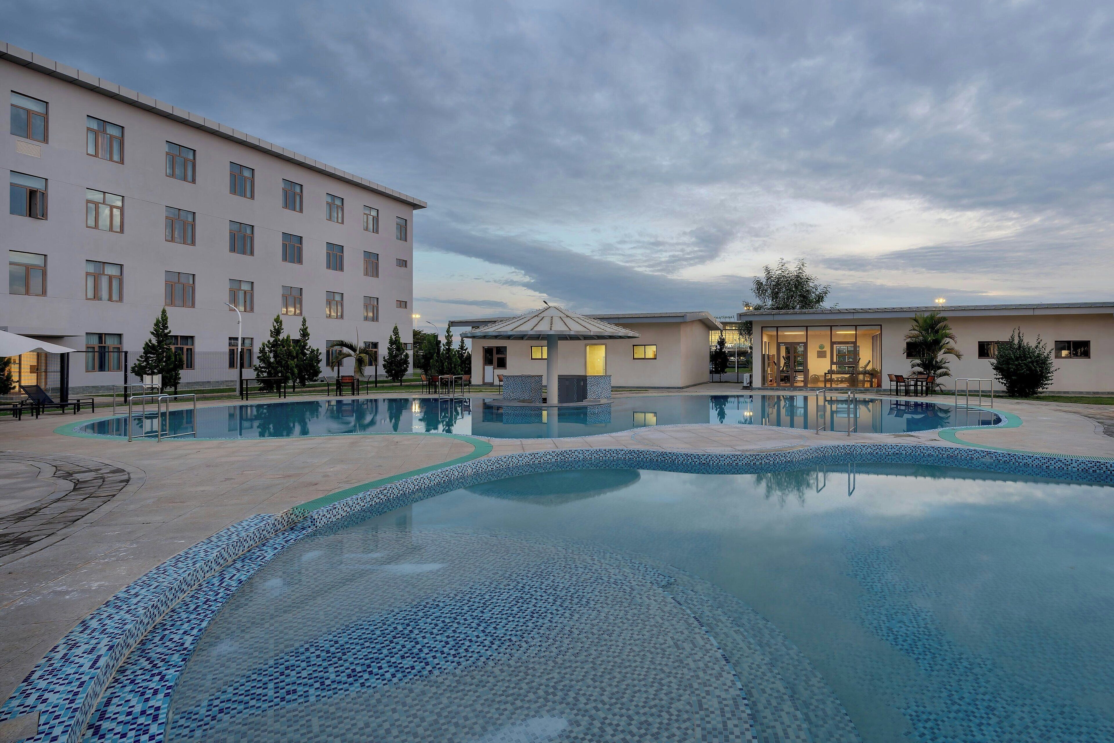 A swimming pool with tiled patterns, surrounded by a building with multiple windows and a small gazebo-like structure.