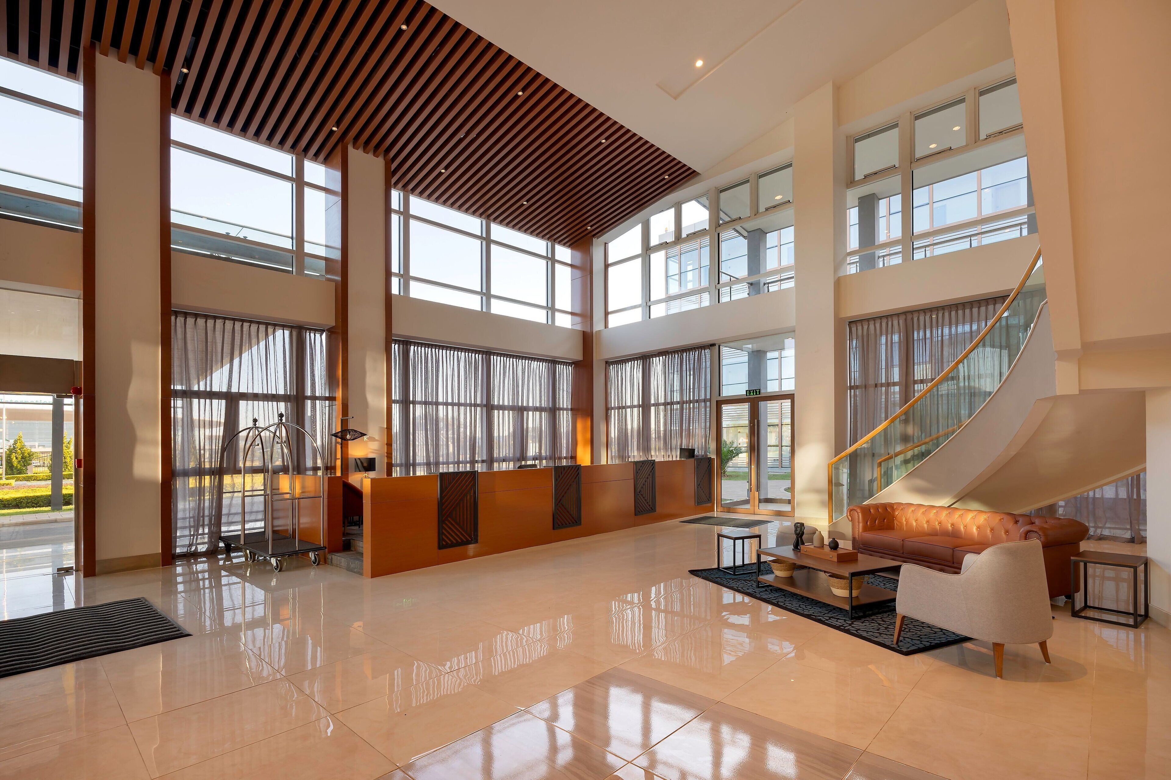 A spacious lobby with a high ceiling, large windows, a staircase, and a reception desk.