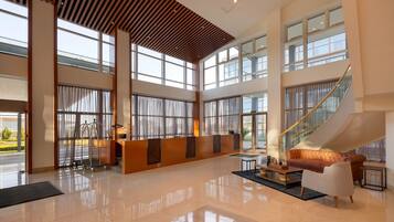 A spacious lobby with a high ceiling, large windows, a staircase, and a reception desk.