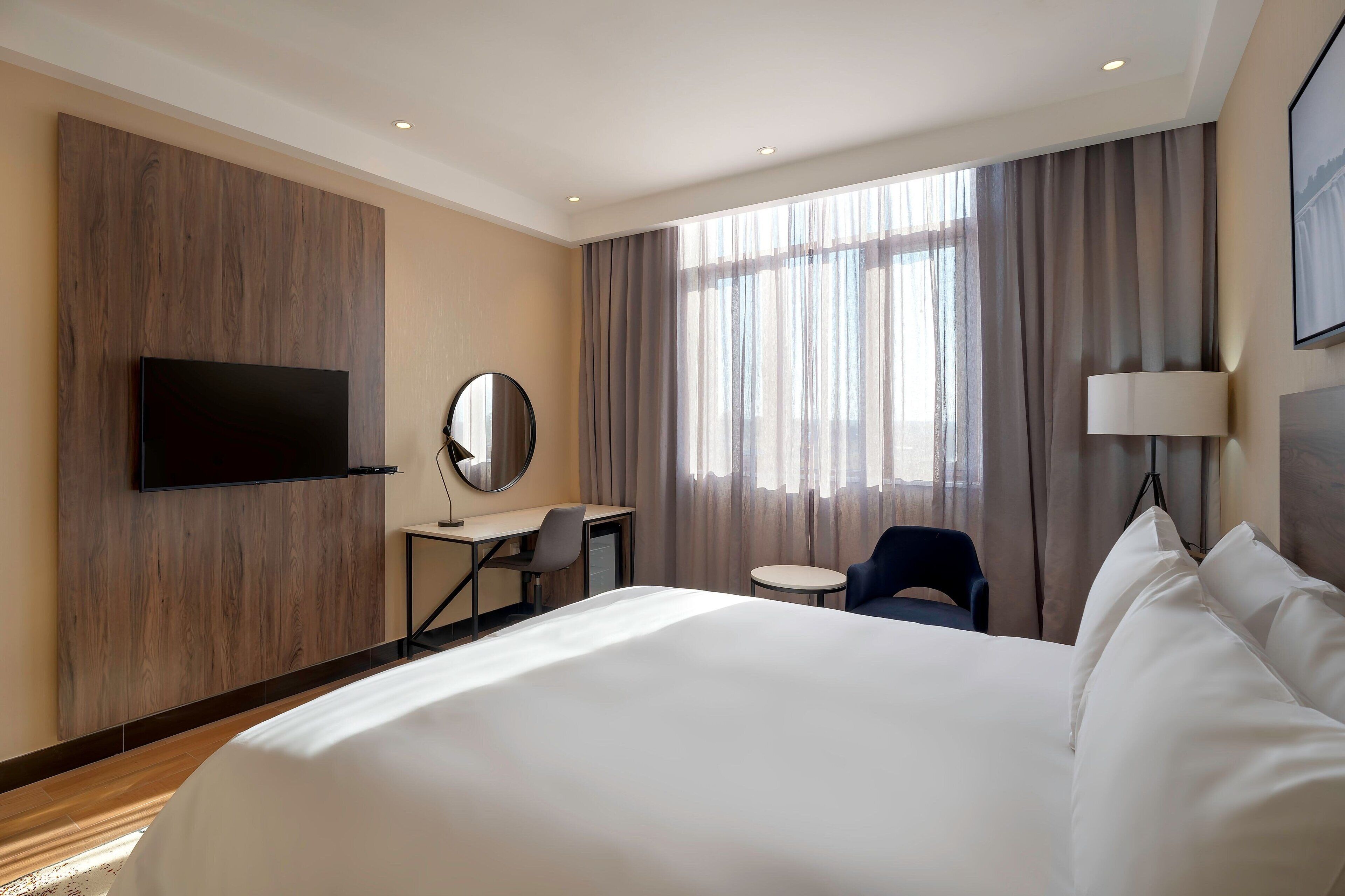 A hotel room with a large bed, a desk, a chair, a TV mounted on the wall, and a window with curtains.