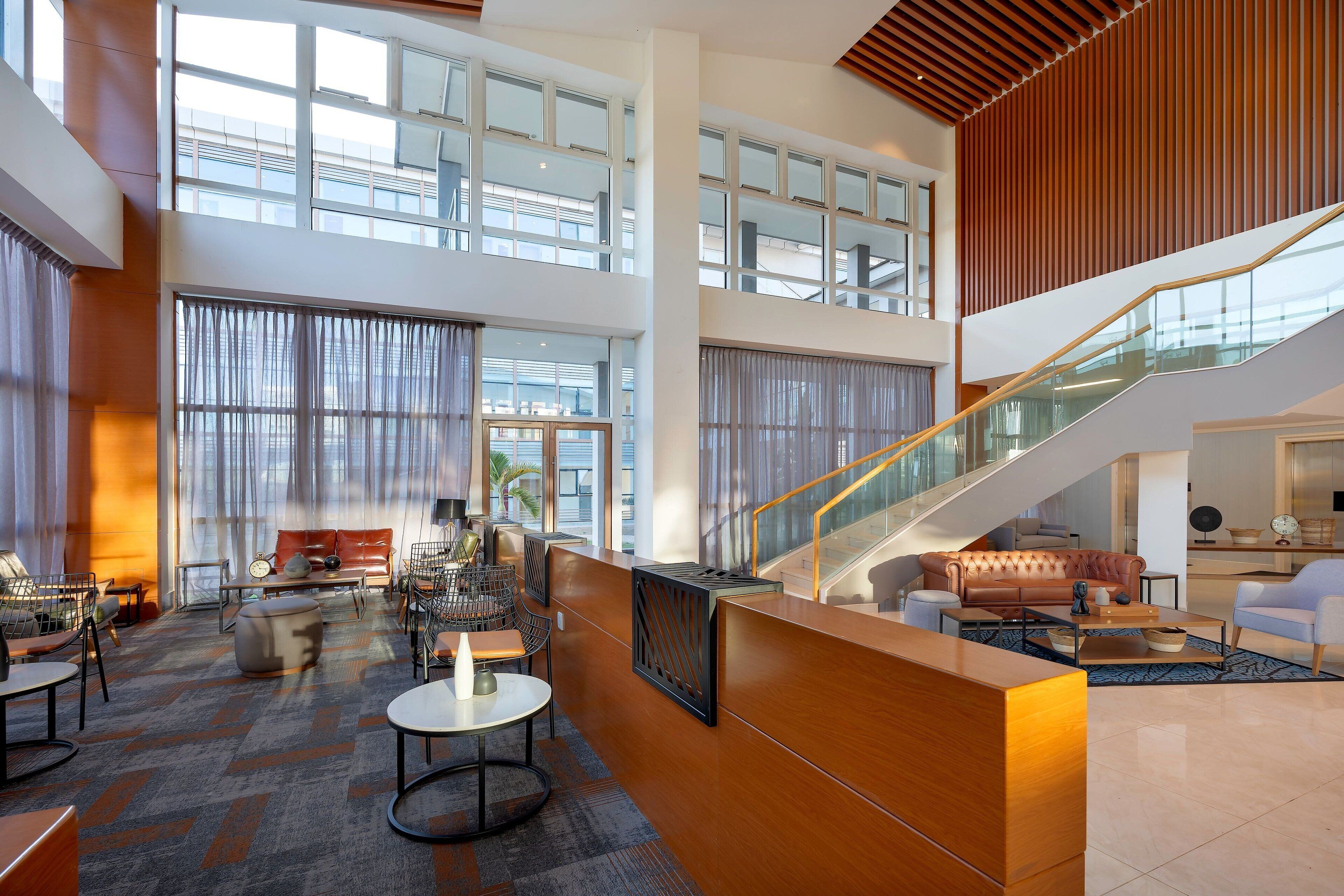 A spacious lobby with a high ceiling, large windows, a staircase, and modern furniture.