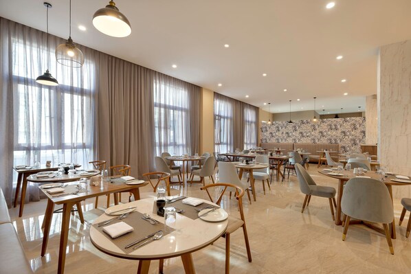 Un amplio comedor con varias mesas y sillas, ventanas grandes con cortinas e iluminación colgante.