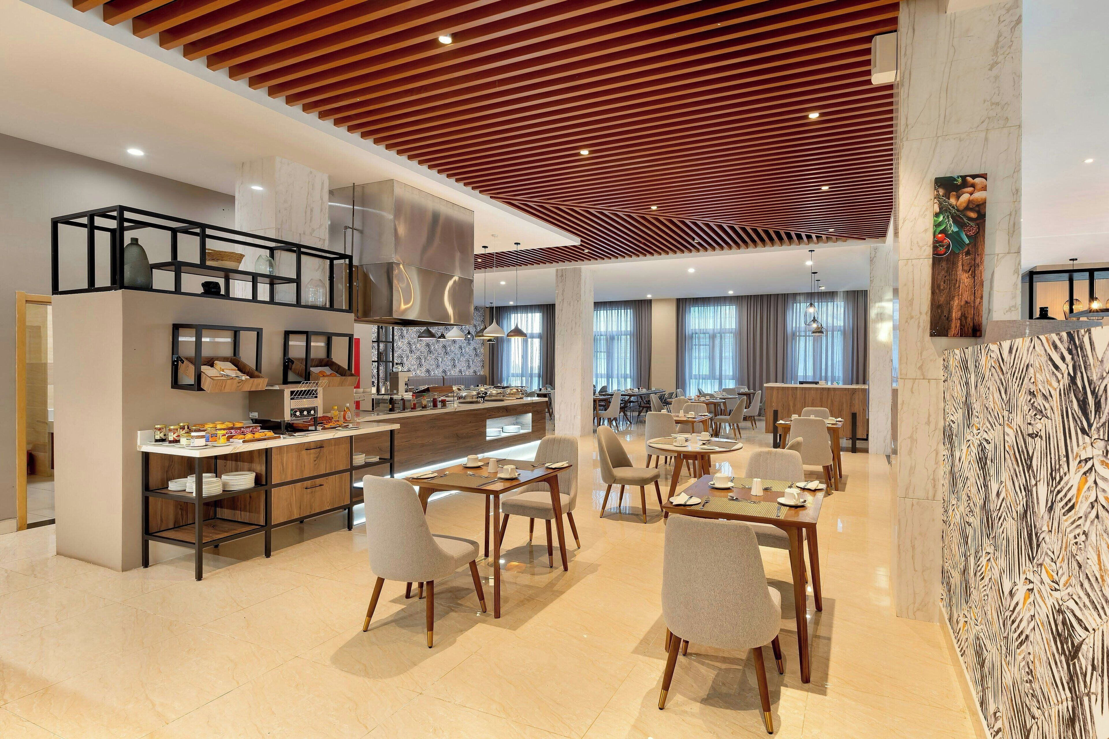 A modern hotel lobby with a kitchen area, dining tables, and chairs.