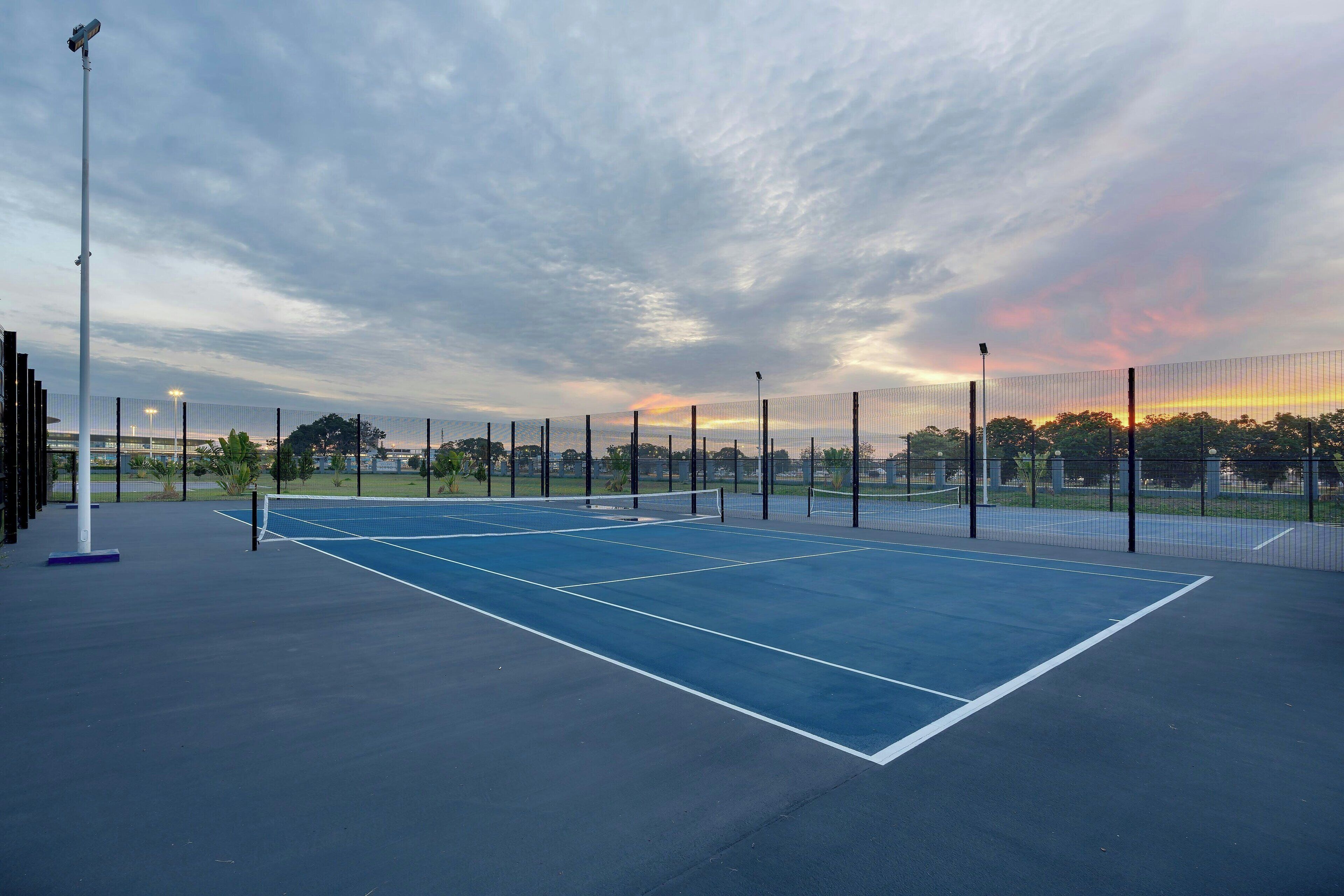 Ein Tennisplatz im Freien mit Netz, umgeben von einem Zaun, unter einem bewölkten Himmel.