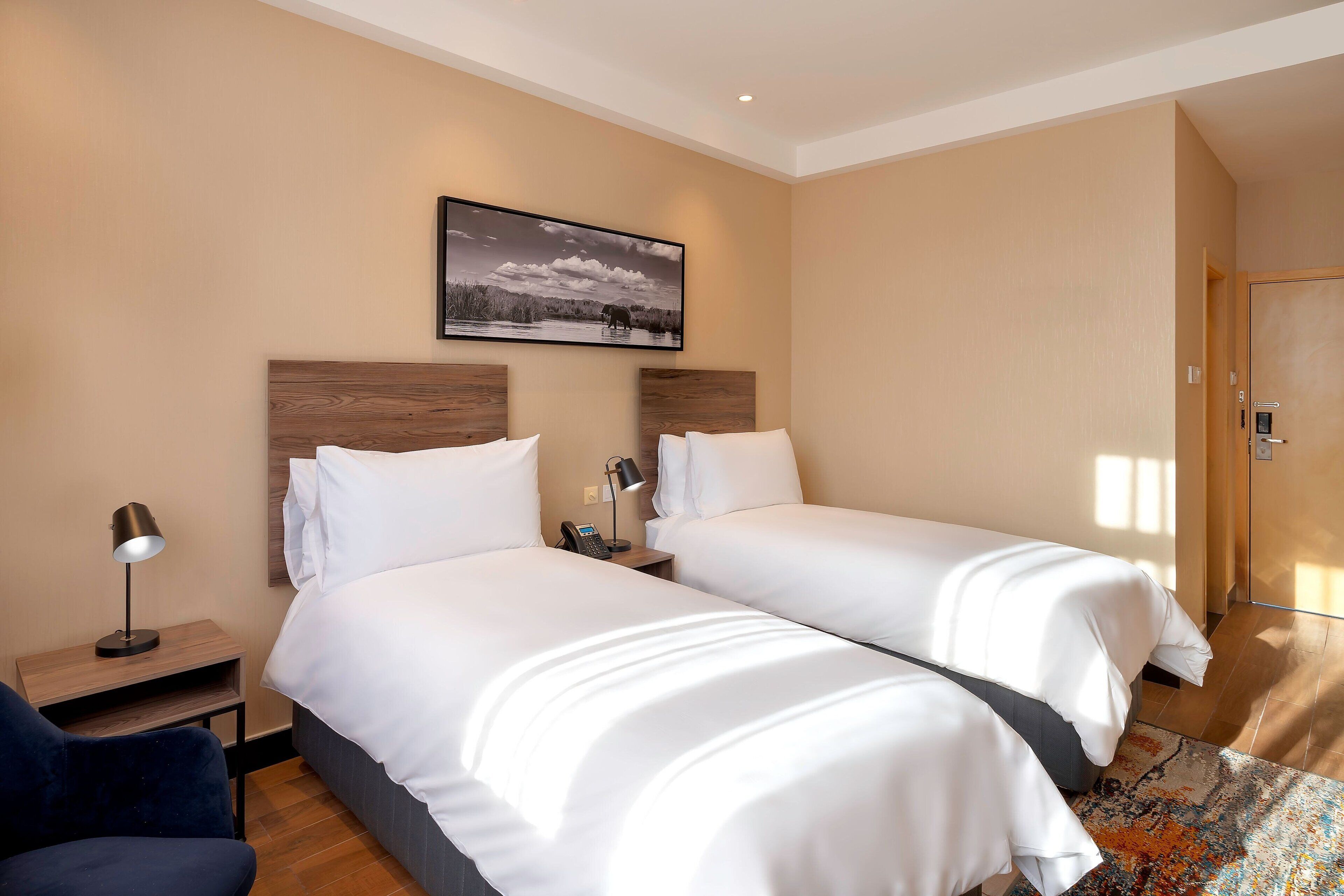 Habitación de hotel con dos camas, una mesita de noche con lámpara, teléfono y un cuadro en la pared.