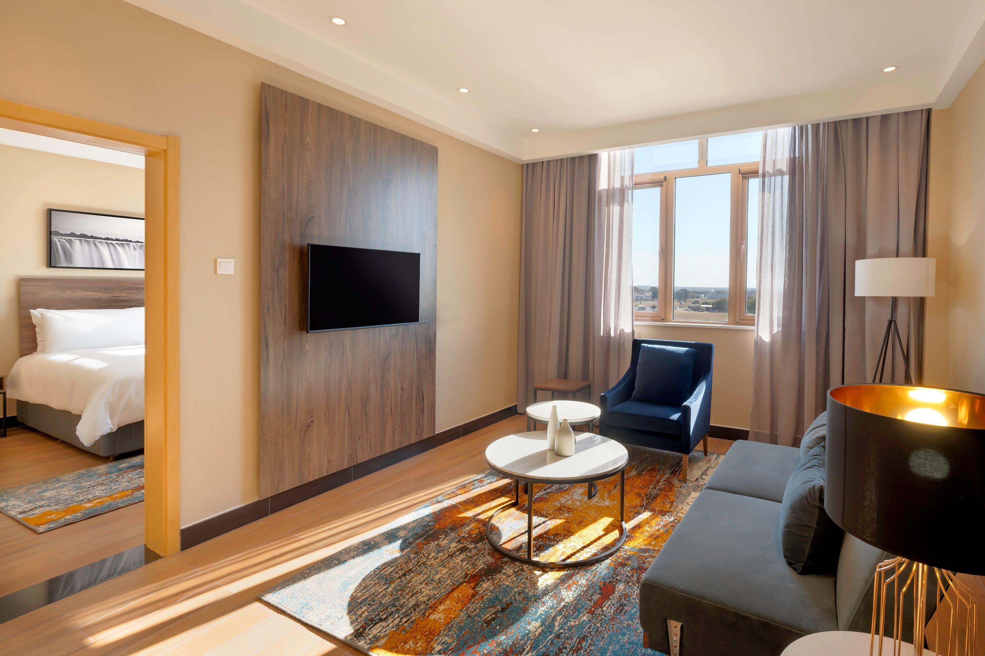 A modern hotel room with a bed, a TV mounted on the wall, a sofa, a small table, and a large window with curtains.