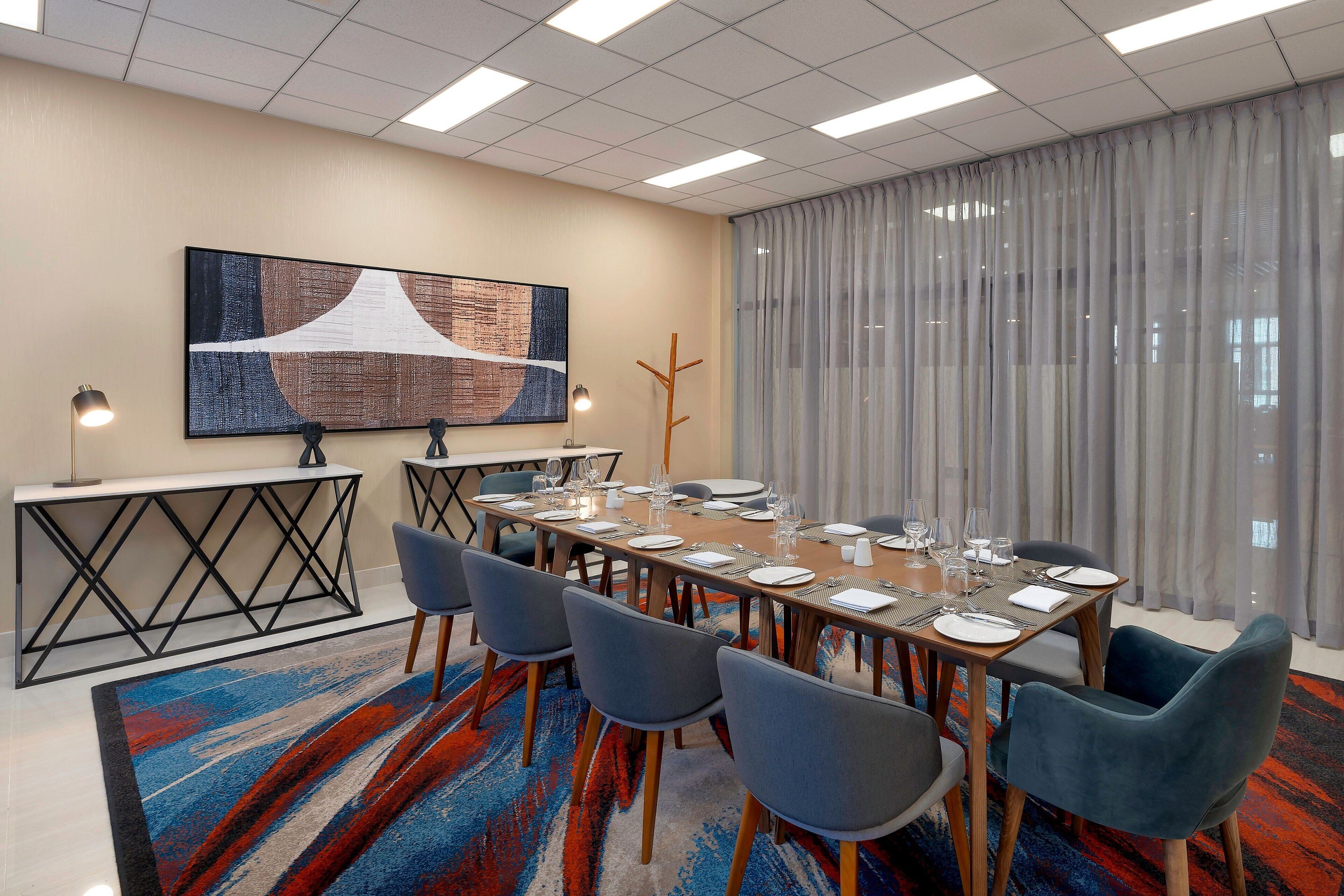 A conference room with a long table set for dining, surrounded by chairs, a decorative wall piece, and a large window with curtains.