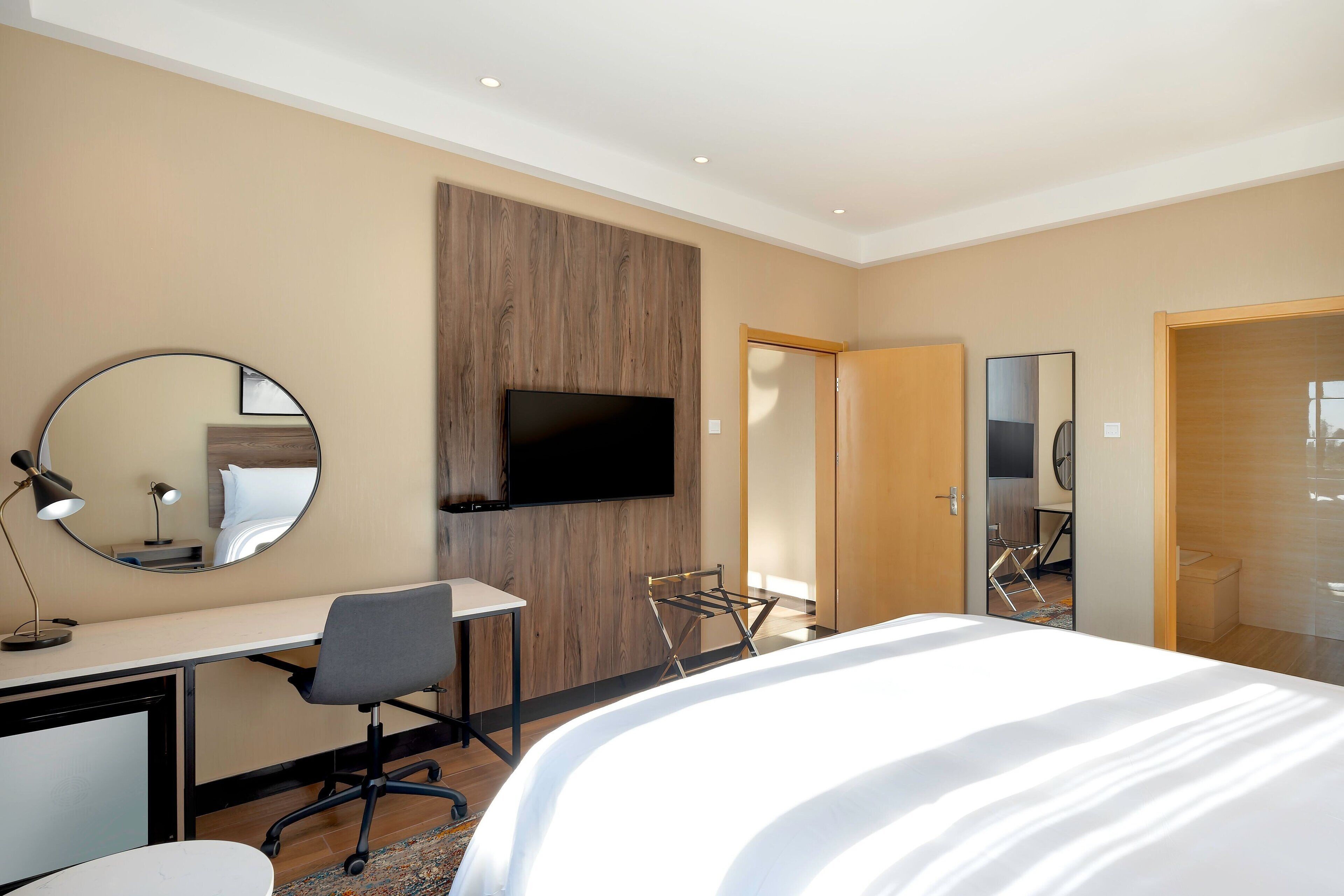 A modern hotel room with a bed, desk, chair, TV, and a mirror.