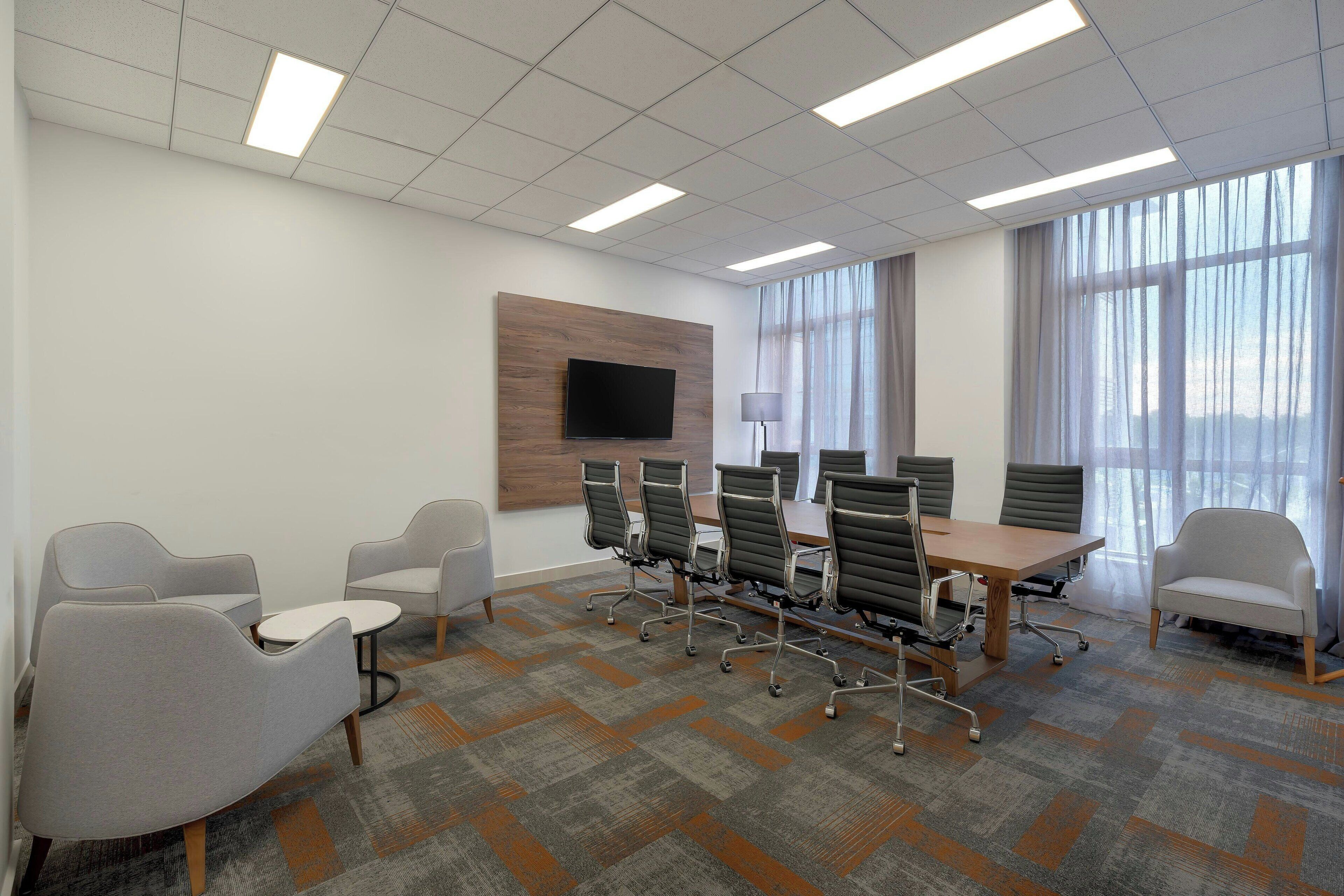 A conference room with a large table, several chairs, a flat-screen TV mounted on the wall, and large windows with sheer curtains.