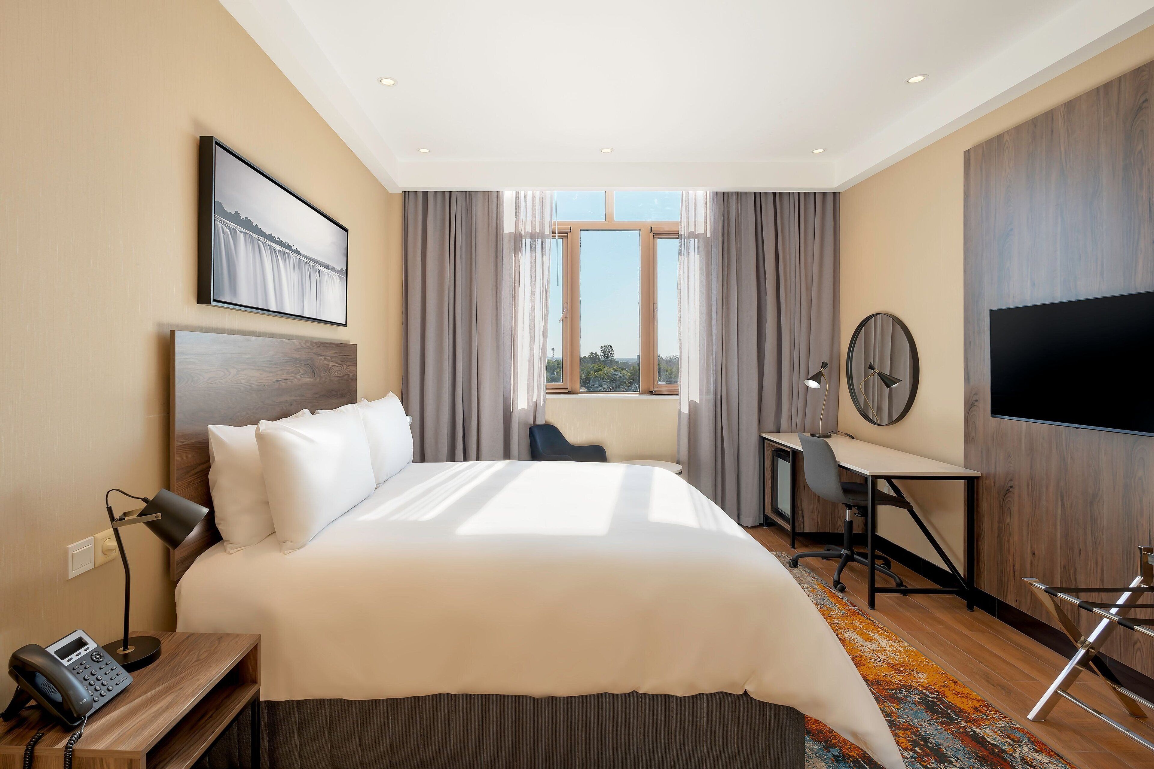 Quarto de hotel moderno com uma cama grande, uma escrivaninha, uma cadeira, uma TV de tela plana e um telefone num criado-mudo.