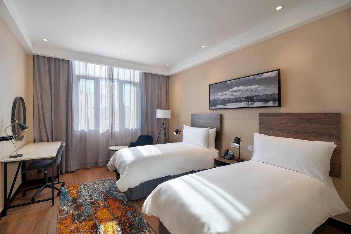 Habitación de hotel con dos camas, un escritorio, una silla, una lámpara y un cuadro en la pared.