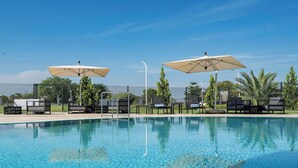 A clear blue swimming pool with lounge chairs and umbrellas, surrounded by greenery and a fence.